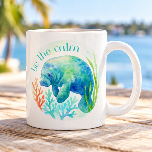 A white ceramic mug with a watercolor-style illustration of a blue-green manatee, coral, and the words “be the calm,” placed on a wooden table with a blurred beach and water background.