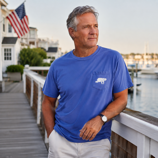 A man wearing a royal blue short-sleeve pocket T-shirt with a white embroidered bear logo on the chest, standing on a wooden dock by the water with houses and an American flag in the background.