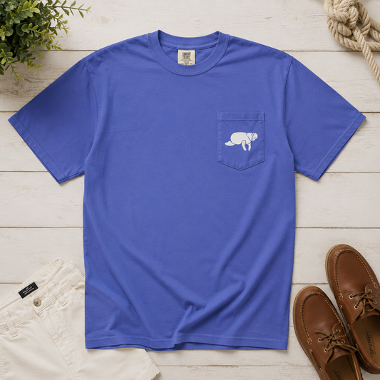 A royal blue short-sleeve pocket t-shirt with a white graphic of a kneeling dog on the chest pocket, styled in a flat lay with white pants, brown boat shoes, rope, and a plant on a white wood plank background.