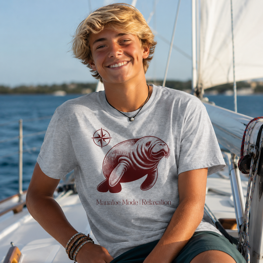 A smiling young man with blonde hair sits on a sailboat wearing a light gray t-shirt with a maroon manatee graphic and the text 'Manatee Mode | Relaxation'.