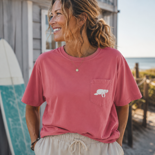 A woman wearing a dusty rose pink short-sleeve t-shirt with a white turtle graphic on the chest pocket, paired with beige drawstring shorts, standing outdoors near a beach boardwalk.
