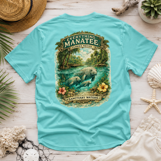 A turquoise short-sleeve t-shirt laid flat on a white wood surface, featuring a large vintage-style graphic on the back with a manatee in a river and the text 'Everything Manatee Florida Native' and 'Sanctuary Waters.' Surrounding props include a straw hat, seashells, a starfish, plant leaves, and a small succulent.