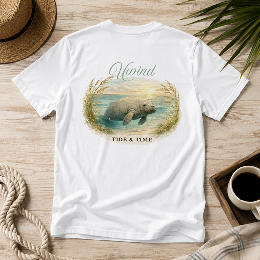A white short-sleeve T-shirt laid flat on a light wood surface, featuring a large back graphic of a manatee in water with the words 'Mivind' above and 'TIDE & TIME' below. The shirt is styled with a straw hat, rope, green leaves, and a cup of coffee.