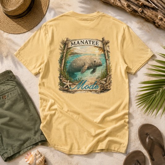 A pale yellow graphic T-shirt laid flat on a light surface with beach-themed props including a straw hat, shorts, sandals, driftwood, and a palm leaf. The shirt has a large front graphic showing a manatee in water with the words “MANATEE Mode” in decorative lettering.