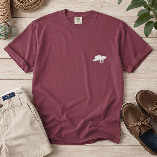 A burgundy short-sleeve pocket T-shirt with a white dog graphic on the chest, styled in a flat lay with khaki shorts, brown boat shoes, rope, and green leaves on a light wood background.
