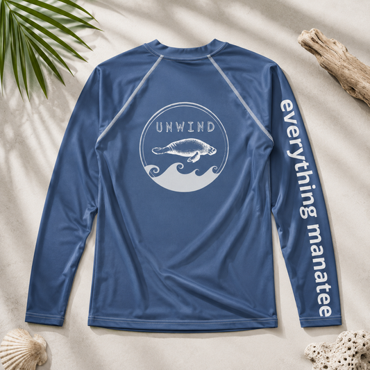 A blue long-sleeve rash guard shirt laid flat on a sandy surface with beach props. The back has a white circular wave graphic with a manatee and the word 'UNWIND' above it, and the right sleeve has 'everything manatee' printed vertically in white.