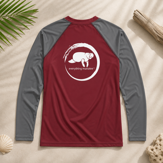 A long-sleeve raglan shirt laid flat on a sandy surface with beach props. The shirt has a burgundy torso and gray sleeves, with a large white chest graphic showing a monkey inside a circular brushstroke and the text 'everything manatee'.