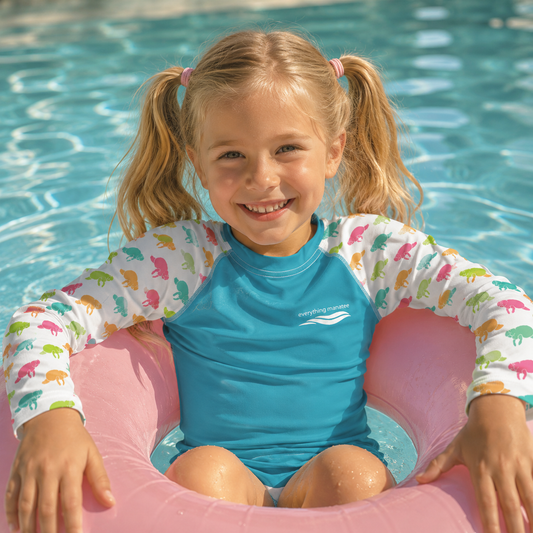 A young girl with blonde pigtails sits in a pink inflatable pool float in a swimming pool, wearing a blue and white long-sleeve swim shirt with a colorful dinosaur print on the sleeves.