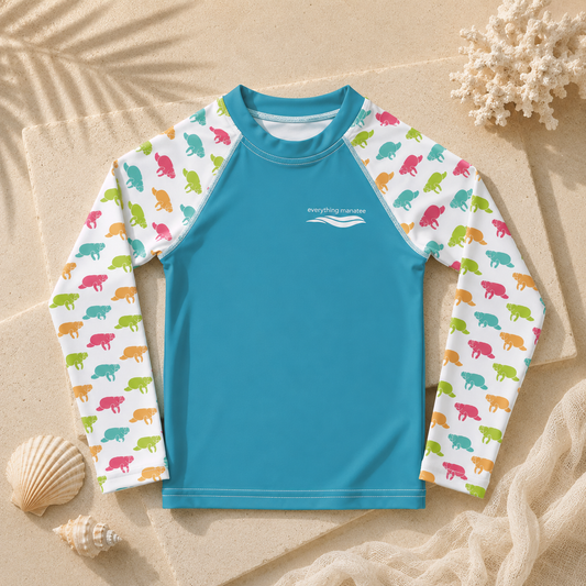 A children's long-sleeve rash guard laid flat on a sandy surface with seashells and coral props. The shirt has a bright turquoise-blue torso and sleeves printed with colorful dinosaur motifs on a white base.