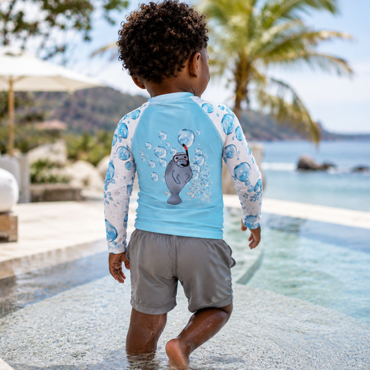 A toddler seen from behind walking into a swimming pool wearing a light blue long-sleeve rash guard with a seal graphic and gray swim shorts, with a poolside resort setting in the background.