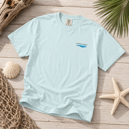 A light blue crewneck t-shirt with a small 'everything natural' wave logo on the left chest, styled in a flat lay on a wooden surface with seashells, a starfish, a net, and driftwood.
