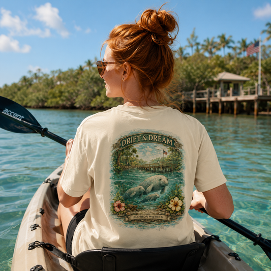 A woman in a kayak seen from behind, wearing a cream-colored t-shirt with a large graphic print of manatees and the text 'DRIFT & DREAM' on the back. She is paddling on turquoise water with tropical islands in the background.
