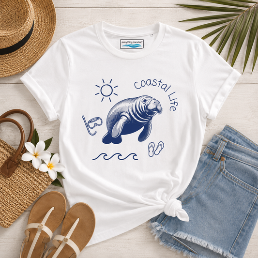 A white graphic t-shirt featuring a navy blue manatee illustration and the words 'coastal life' with beach-themed props including denim shorts, a straw hat, sandals, and flowers on a light wood background.