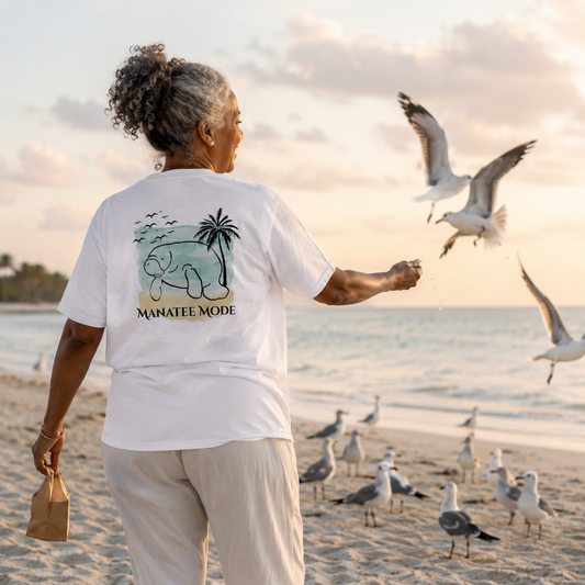 An older woman stands on a beach at sunset facing the ocean, wearing a white graphic T-shirt with a teal elephant and palm tree design and the words 'MANATEE MODE' on the back, while feeding seagulls.
