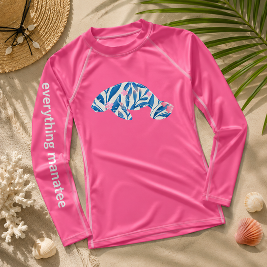 A bright pink long-sleeve rash guard laid flat on a sandy beach background with seashells, a straw hat, and palm leaves. The shirt has a blue and white tropical leaf patterned seal graphic on the chest and 'everything manatee' printed in white down one sleeve.