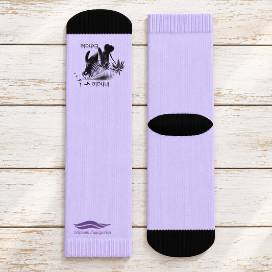 Two light purple socks displayed flat on a white wood plank background. One sock shows the front with a black toe cap, black printed graphic, and purple logo near the foot; the other shows the back with a black heel cutout and black toe cap.