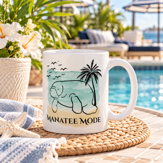 A white ceramic mug with a manatee beach graphic and the text 'MANATEE MODE' sits on a woven placemat by a poolside table. The background shows a blurred swimming pool, lounge chairs, palm trees, and white flowers.