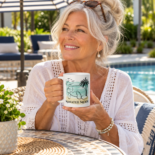 A smiling older woman sits at an outdoor patio table by a pool, holding a white ceramic mug with a teal and black tropical graphic and the words “MANATEE MODE.” She wears a white crochet top and sunglasses rest on her head.