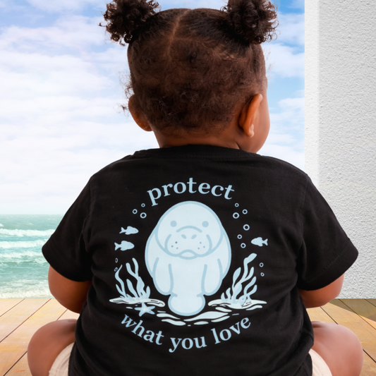 Child wearing a black t-shirt with an environmental message and cartoon character, sitting by a window with a view of the ocean.
