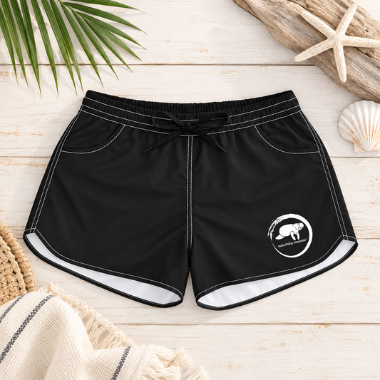 Black swim shorts with white contrast stitching, a black drawstring waist, and a white circular logo on the lower left leg, displayed in a flat-lay arrangement on a light wood background with palm leaves, driftwood, a seashell, and a woven towel.