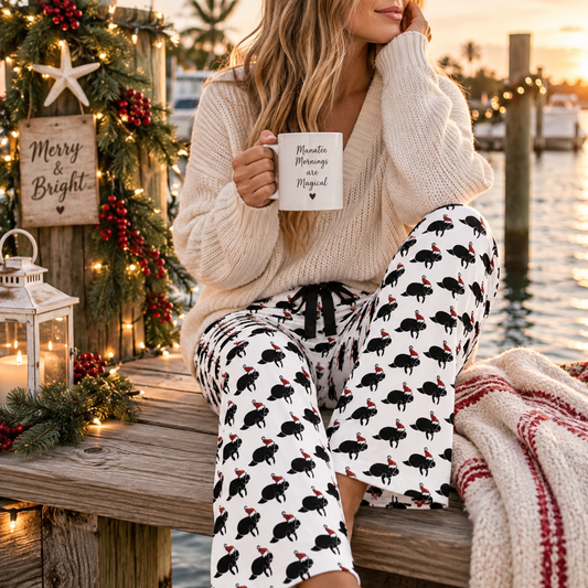 A woman sitting on a wooden dock by the water at sunset, wearing a chunky cream knit sweater and white pajama pants printed with black flamingos and small red accents. She holds a mug that reads 'Merry Night, Merry Christmas.