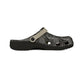 A single clog-style sandal shown in side profile on a plain white background. The upper is black with a light gray leaf pattern, and the strap is a light beige/tan color with a dark snap button.