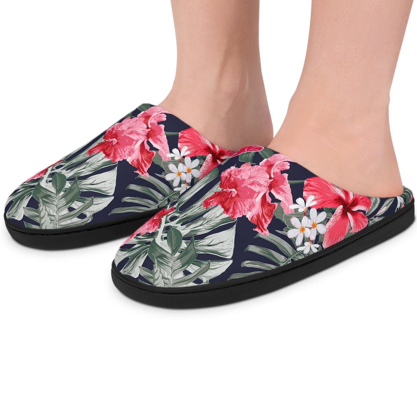 A pair of tropical print slip-on slippers worn on feet, shown against a plain white background. The slippers feature a navy blue base with pink hibiscus flowers, green palm leaves, and small white daisies.