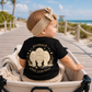 A baby wearing a beige bow headband and a black t-shirt with a beige graphic of three manatees and the text 'protect what you love', sitting in a stroller on a wooden boardwalk by the beach.