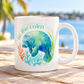 A white ceramic mug with a watercolor-style illustration of a blue-green manatee, coral, and the words “be the calm,” placed on a wooden table with a blurred beach and water background.