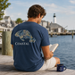 A man sitting on a wooden dock by the water reads a book, wearing a navy blue t-shirt with a manatee graphic and the word 'COASTAL' printed on the back. A matching navy mug with a white anchor sits next to him.