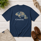 A navy blue short-sleeve t-shirt laid flat on a white wood plank surface, featuring a camouflage-patterned manatee graphic and the word 'COASTAL' in white. The shirt is styled with beige pants, brown boat shoes, a rope, and green leaves.
