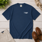 A navy blue crewneck t-shirt with a small white chest graphic reading 'everything normal' and a wave design, styled in a flat lay on a light wood background with khaki pants, brown boat shoes, greenery, and a rope.