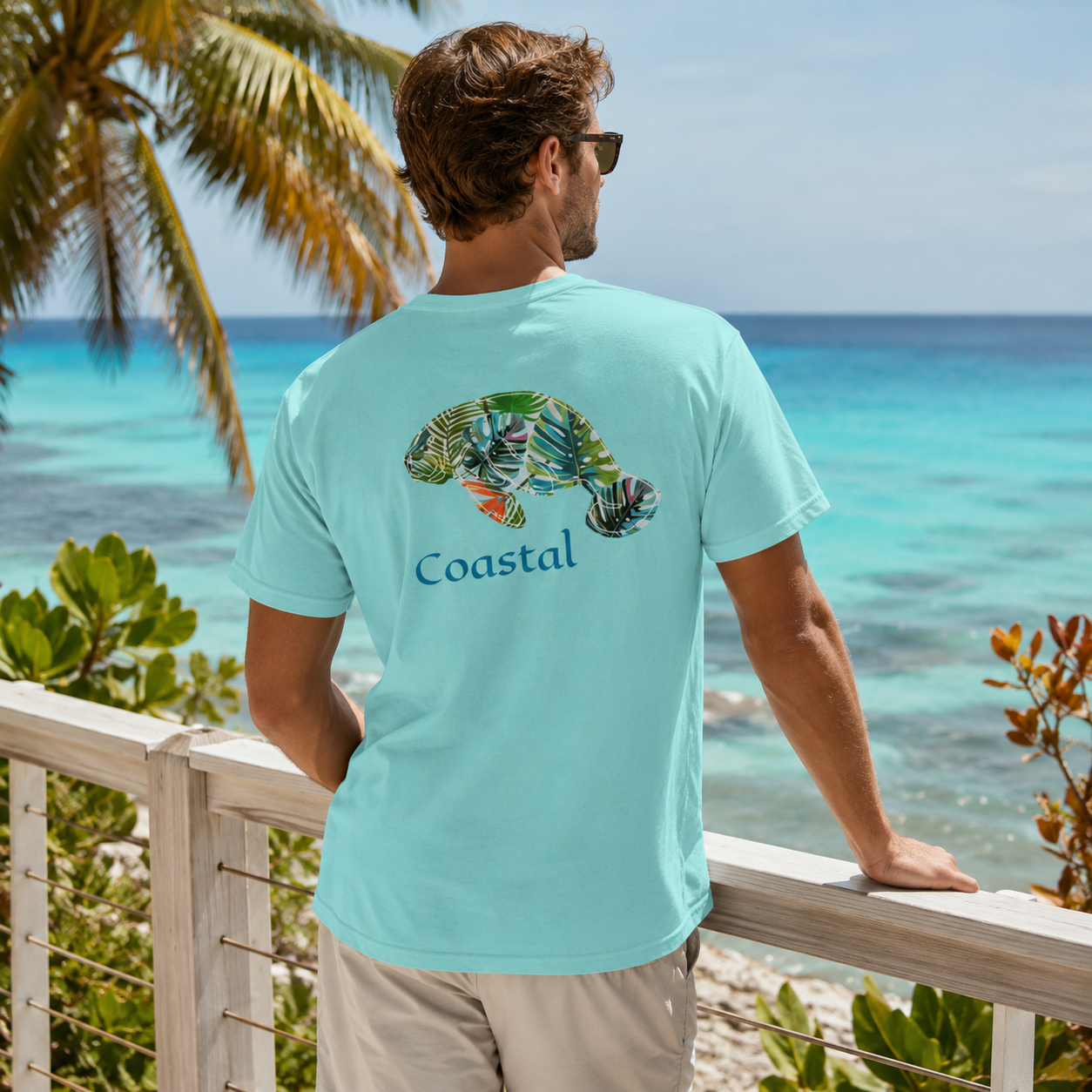 A man wearing a light blue-green t-shirt with a colorful turtle graphic and the word 'Coastal' printed on the back, standing on a balcony overlooking a tropical ocean.