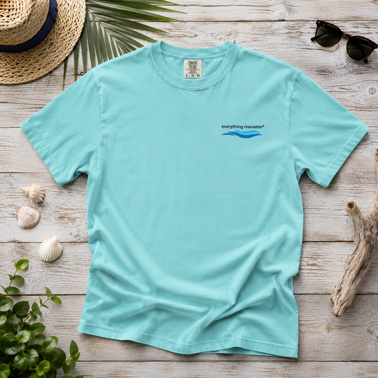 A light aqua blue short-sleeve t-shirt with a small 'everything matters*' logo printed on the left chest, styled in a flat-lay on a whitewashed wooden surface with a straw hat, sunglasses, driftwood, shells, and green leaves.