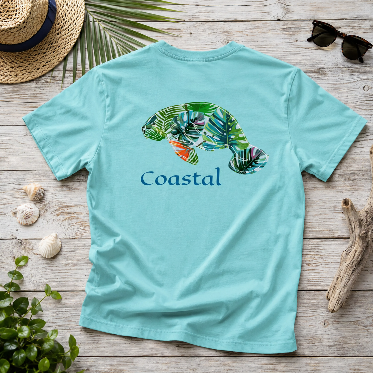A light aqua blue short-sleeve t-shirt with a graphic of a green tropical leafy sea turtle and the word 'Coastal' printed on the back, styled in a flat-lay on a rustic wooden background with a straw hat, sunglasses, driftwood, and seashells.