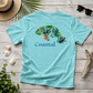 A light aqua blue short-sleeve t-shirt with a graphic of a green tropical leafy sea turtle and the word 'Coastal' printed on the back, styled in a flat-lay on a rustic wooden background with a straw hat, sunglasses, driftwood, and seashells.