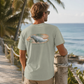 A man wearing a sage green t-shirt with a graphic of a manatee on a beach, photographed from behind as he looks out towards the ocean.