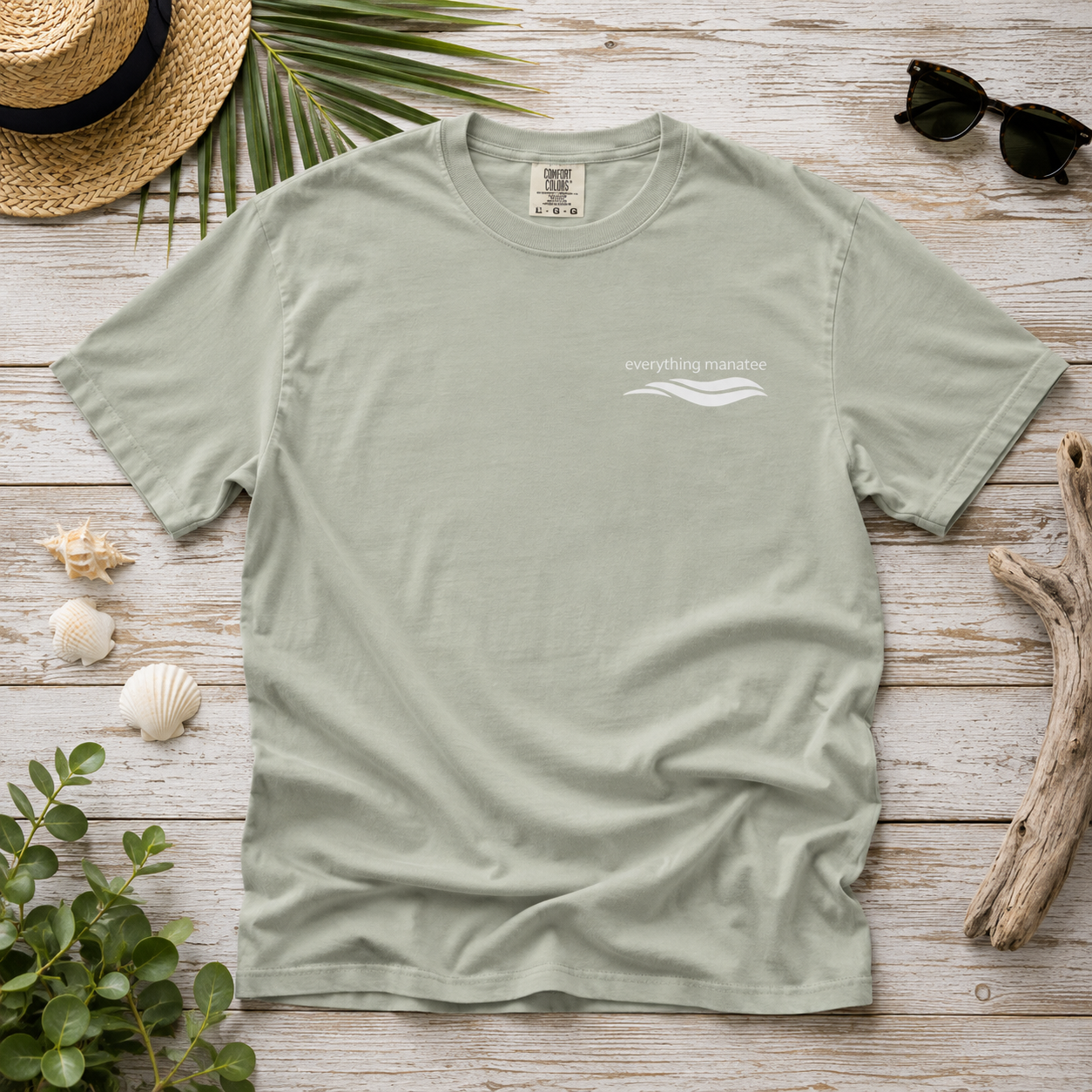 A sage green crewneck t-shirt with a small white graphic on the left chest reading 'everything normal' with a wave design. The t-shirt is displayed in a flat-lay arrangement on a light wood background, surrounded by a straw hat, palm leaves, sunglasses, seashells, driftwood, and green leaves.