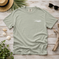 A sage green crewneck t-shirt with a small white graphic on the left chest reading 'everything normal' with a wave design. The t-shirt is displayed in a flat-lay arrangement on a light wood background, surrounded by a straw hat, palm leaves, sunglasses, seashells, driftwood, and green leaves.