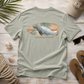 A sage green t-shirt laid flat on a rustic white wood background, featuring a graphic of a manatee on a surfboard with the text 'everything manatee'. The shirt is styled with beach-themed props including a baseball cap, sunglasses, flip-flops, rope, and seashells.