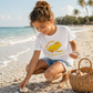 A young girl crouches on a sandy beach next to the ocean, wearing a white graphic t-shirt that reads 'Sunshine & Seashells' with a yellow turtle design. She is holding a seashell and interacting with shells on the sand, with a woven basket nearby.