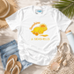 A white short-sleeve graphic T-shirt laid flat on a light wood surface with beach-themed props including denim shorts, sandals, a straw hat, palm leaves, a net bag, and a bottle. The shirt has a yellow sun-and-seashell graphic with the text 'Sunshine & Seashells.