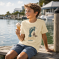 A young boy sitting on a wooden dock by the water, wearing a cream-colored graphic t-shirt with a blue manatee design and the text 'KICK INTO MANATEE MODE and Relax'. He is holding an ice cream cone.