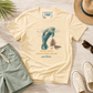 A cream-colored graphic t-shirt featuring a manatee illustration and the text 'KICK INTO MANATEE MODE and Relax' in blue and brown, styled in a flat lay with sage green shorts, white sneakers, a straw hat, sunglasses, and a palm leaf on a light wood background.