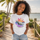 A smiling young girl with curly hair stands on a sandy beach path wearing a white graphic T-shirt and denim shorts. The shirt features a colorful manatee and sun design with the text 'everything manatee'.
