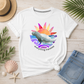 A white graphic t-shirt featuring a colorful manatee illustration and the text 'everything manatee' on the front, styled in a flat-lay with a straw hat, denim shorts, and beach-themed props on a light wood background.