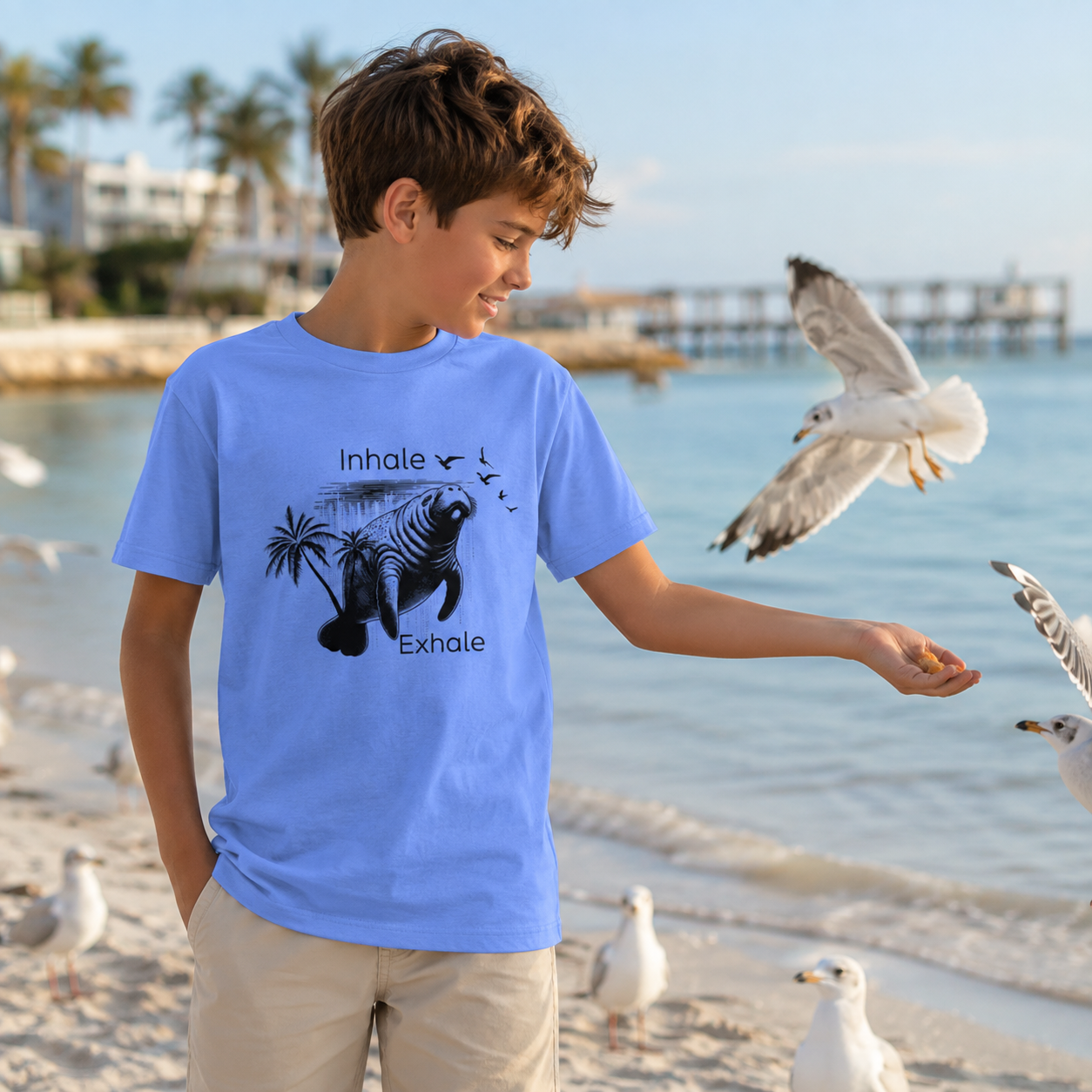 A boy wearing a light blue graphic t-shirt stands on a beach, reaching out to a seagull. The t-shirt features a black graphic of a seal and palm tree with the words 'Inhale' and 'Exhale'.