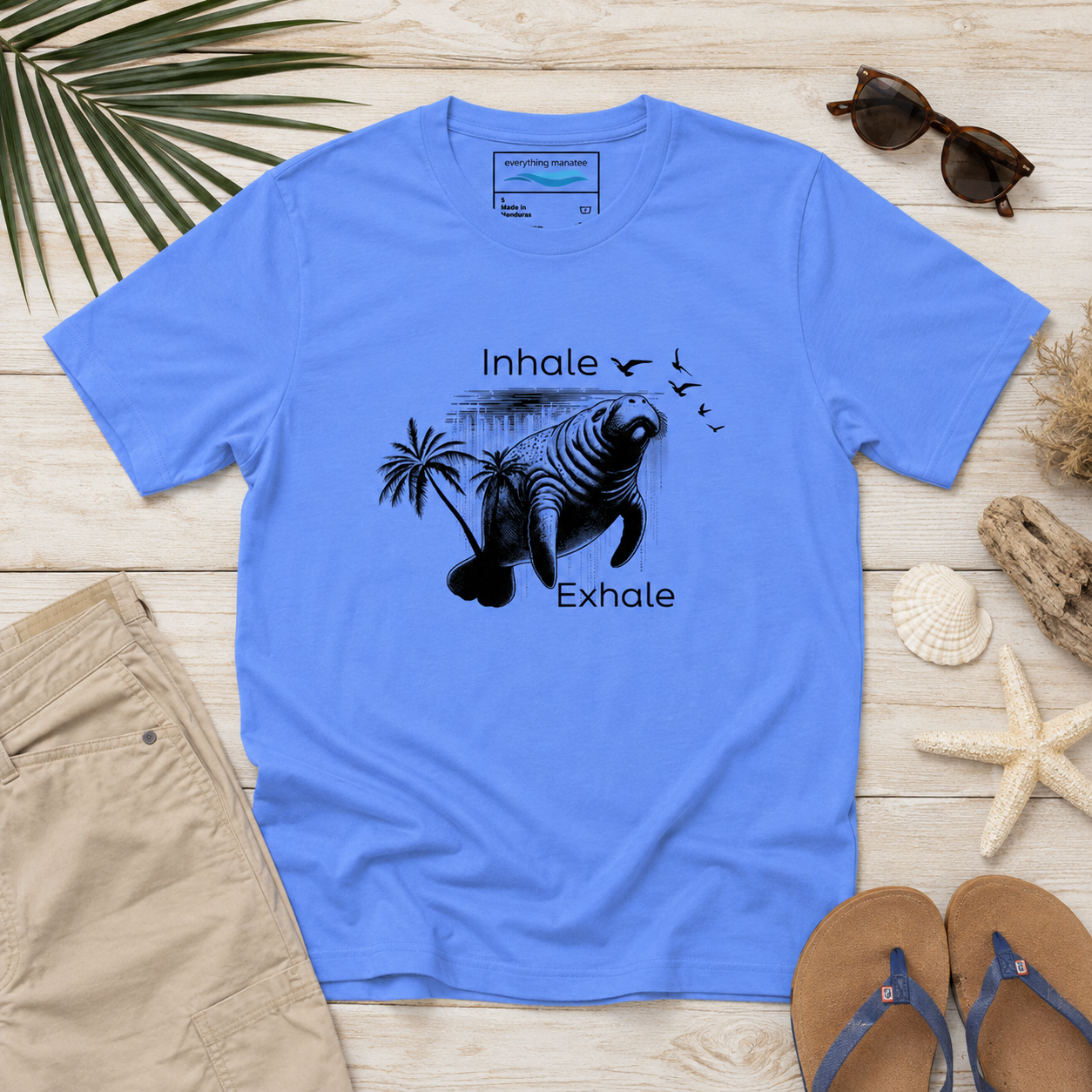 A light blue graphic t-shirt laid flat on a white wood background, styled with sunglasses, khaki shorts, a palm leaf, shells, driftwood, and flip-flops. The shirt features a black graphic of a manatee with palm trees and the words 'Inhale' and 'Exhale'.