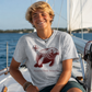 A smiling young man with blonde hair sits on a sailboat wearing a light gray t-shirt with a maroon manatee graphic and the text 'Manatee Mode | Relaxation'.