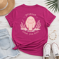 A bright pink t-shirt with a cute pink manatee graphic and the text 'protect what you love' on the back, styled in a flat lay with denim shorts, white sneakers, a straw hat, and a gold bracelet on a white wood background.